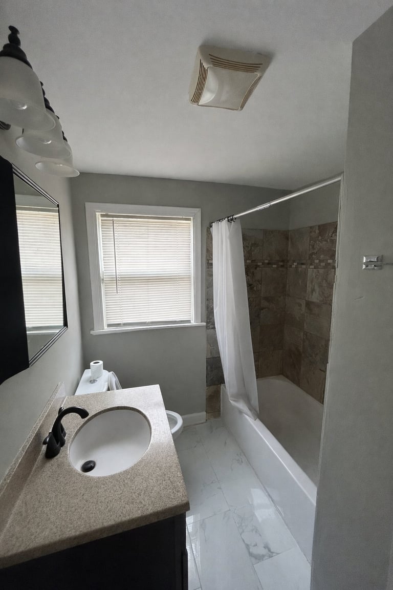 Modern bathroom with white toilet, gray vanity countertop, shower with white curtain, window with blinds, and tiled flooring
