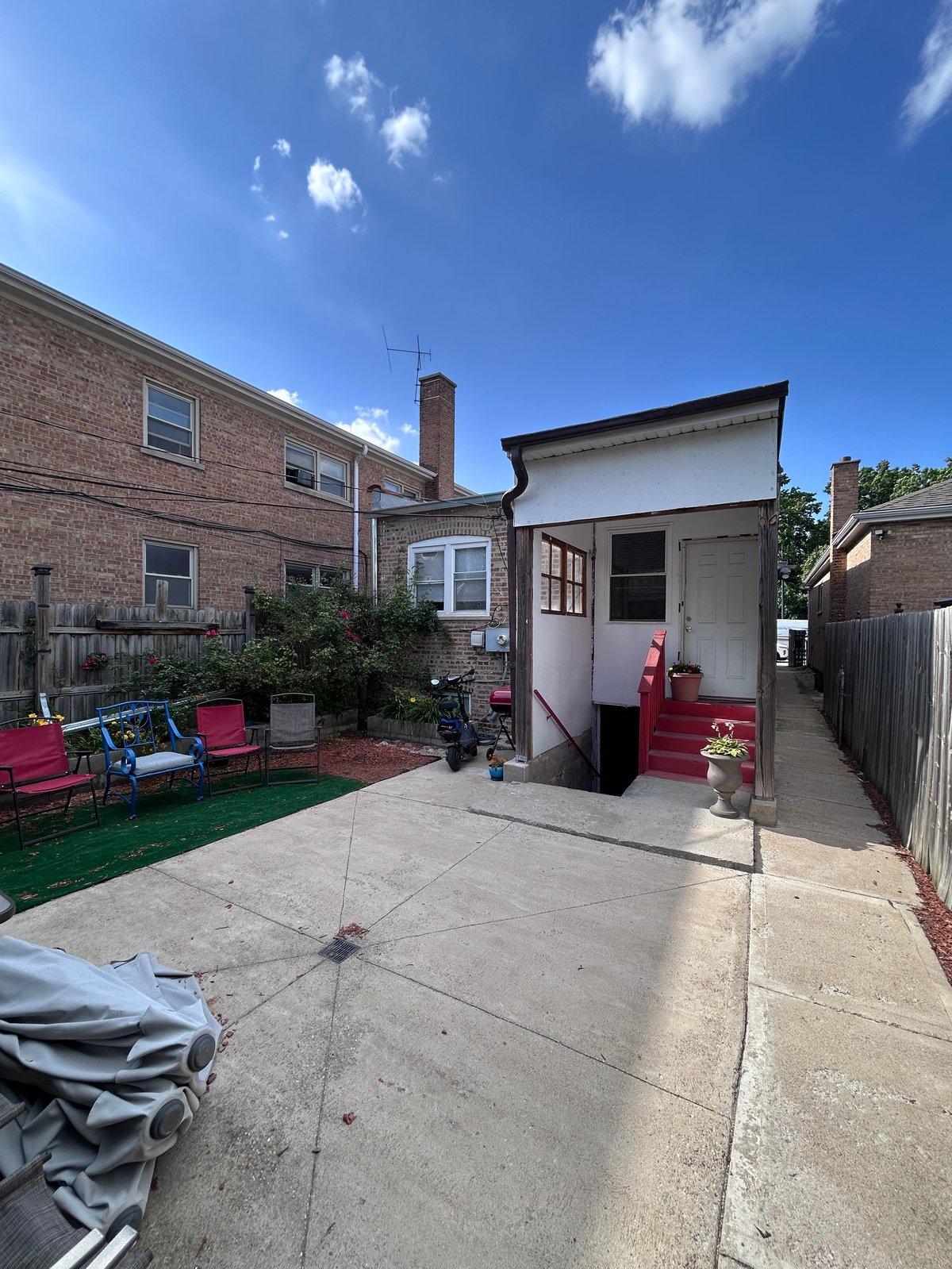 Backyard of brick house with patio, white garage structure, pink seating, and green artificial turf under clear blue sky