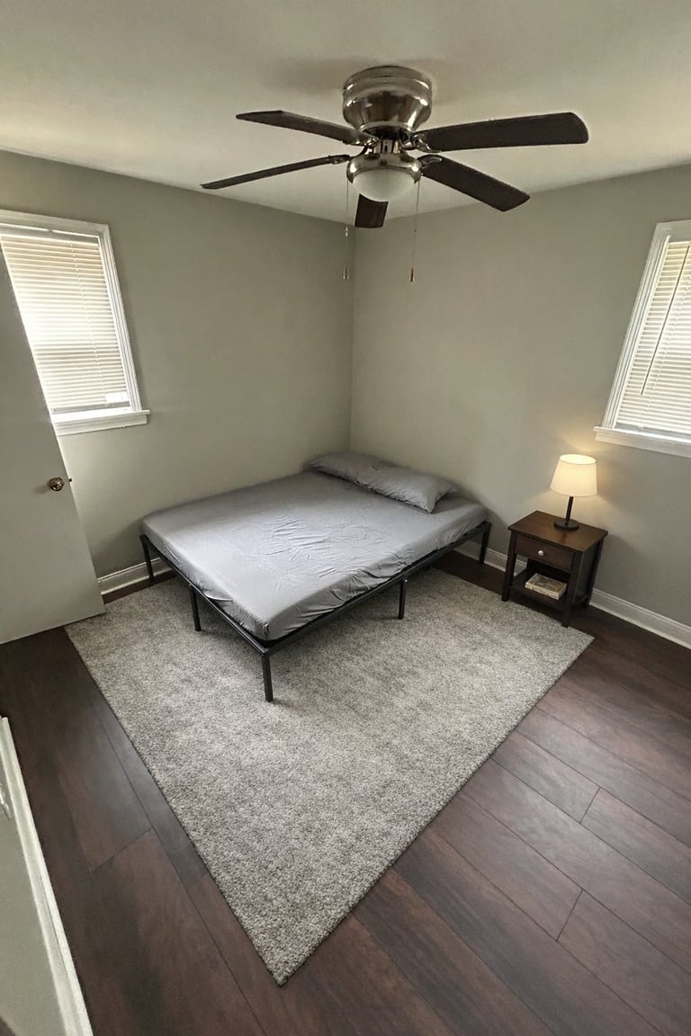 Minimalist bedroom with a simple bed, ceiling fan, wooden nightstand with lamp, and two windows with shutters