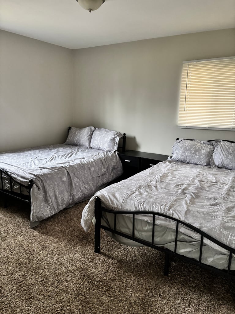 Two twin beds with gray bedding in a bedroom with beige walls, carpet flooring, and a window with blinds