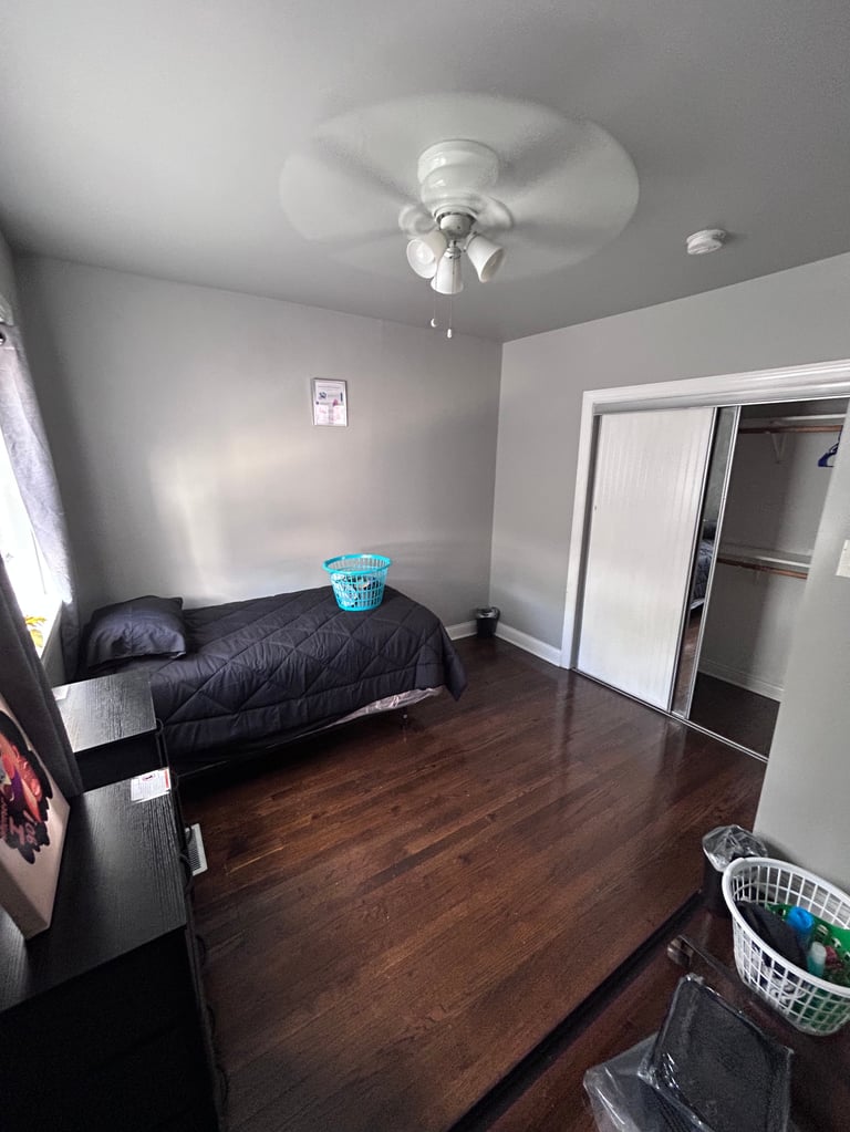 Spacious bedroom with wooden floor, black bed, ceiling fan, white walls, closet, and minimal furnishings