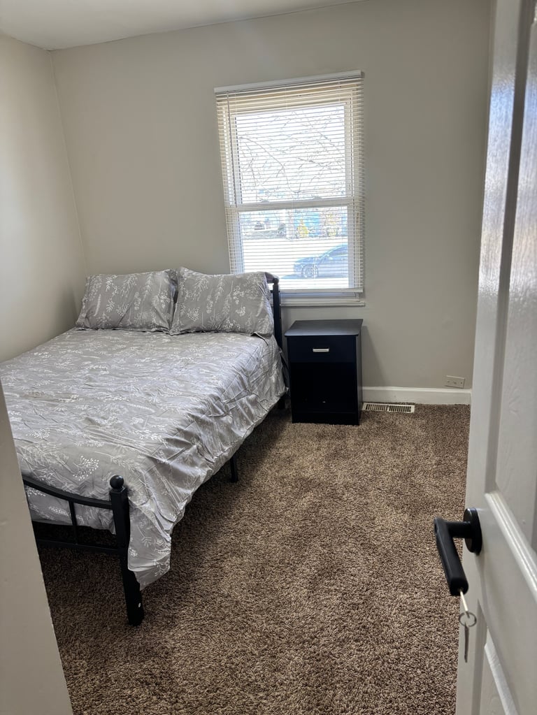 Small bedroom with a queen bed, gray patterned bedding, black nightstand, window with blinds, and beige walls
