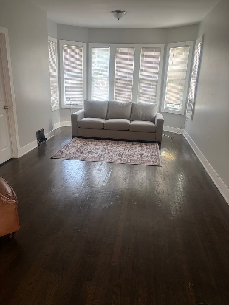 Bright living room with gray sofa, dark hardwood floors, area rug, and large bay window with shutters