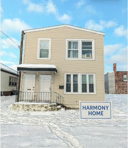 Two-story beige house with white trim and blue Harmony Home sign on snowy ground under clear sky
