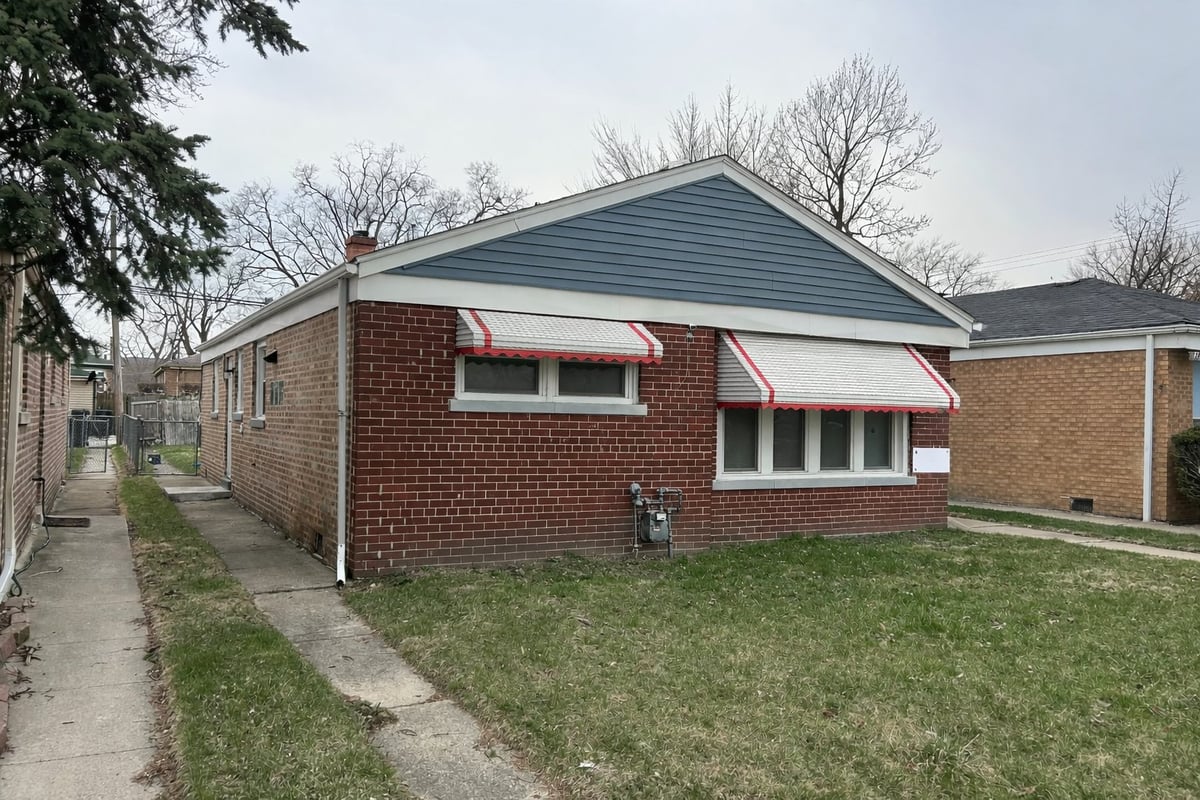 Single-story brick house with blue-grey roof, green lawn, concrete sidewalk, and bare trees in background