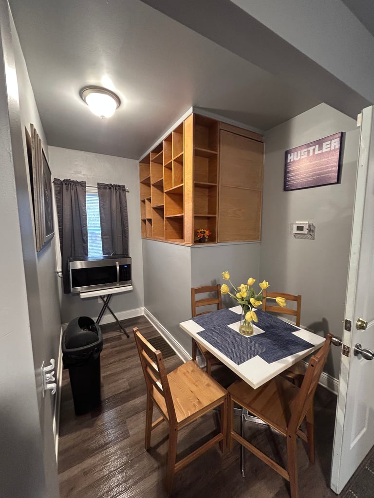 Small dining nook with wooden table, chairs, and built-in shelving above, beside a desk area with dark wood flooring