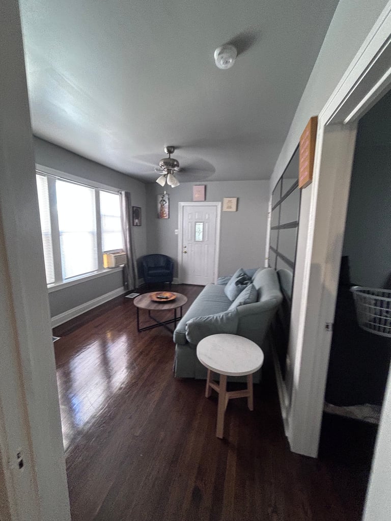 A bright modern living room with gray walls, hardwood flooring, gray couch, white side table, and large windows providing natural light