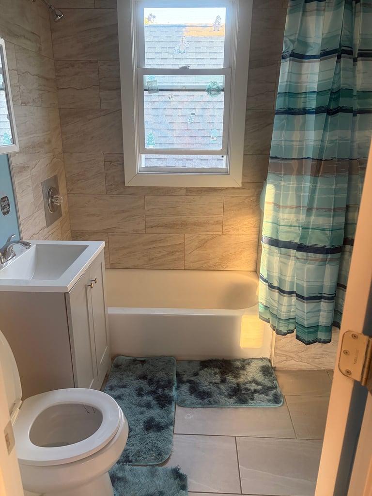 Modern bathroom with wood-look tile, white toilet, soaking tub, green plaid shower curtain, and window overlooking water