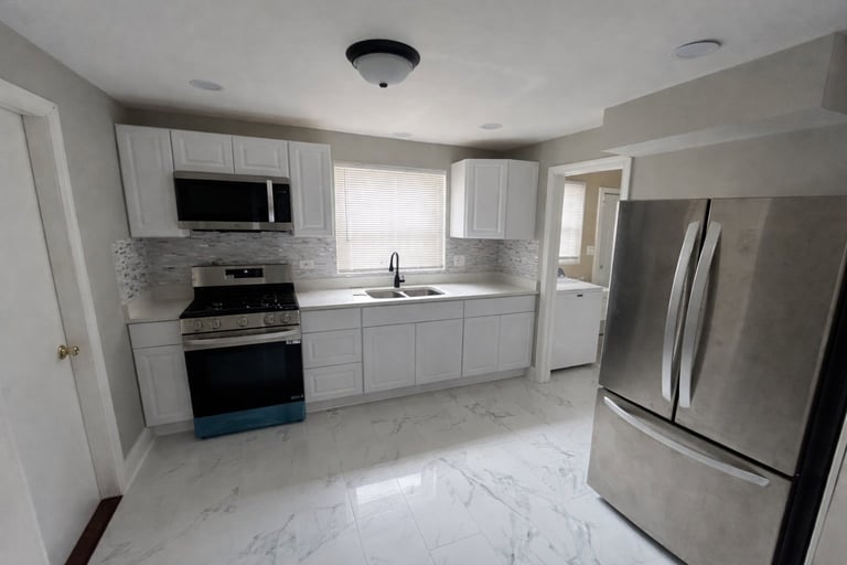 Modern white kitchen with stainless steel appliances, marble countertops, and bright natural light from window