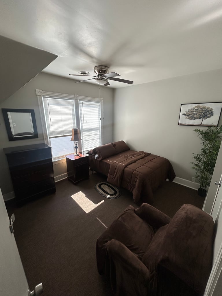 Bedroom with ceiling fan, brown bed, armchair, dark furniture, and glass doors with blinds overlooking natural light